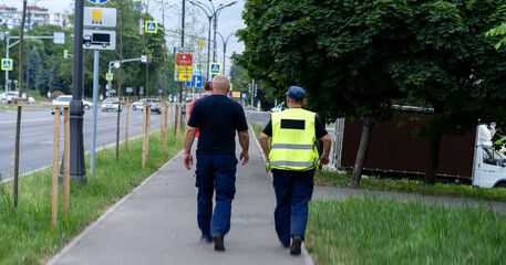 Two uniformed men walking on a city sidewalk near busy street surrounded by greenery during daylight