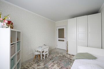 bright room featuring a white wardrobe, bed, shelf, and a small table with chairs. A patterned rug adds warmth to the minimalist space. The walls are light textured.