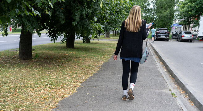 Young woman walking on a city sidewalk surrounded by trees during the day with casual attire and relaxed demeanor - Powered by Adobe