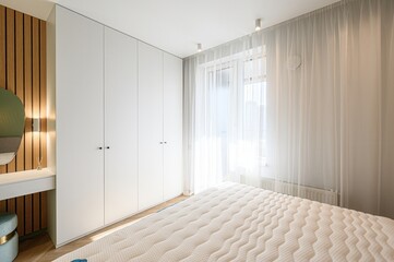bright bedroom features a white bed, large white wardrobe, sheer curtains over a window, a wooden...