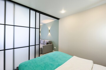 Cozy bedroom with a frosted glass partition, separating a living area with a gray sofa and turquoise curtains. A bright turquoise bedspread adds a pop of color