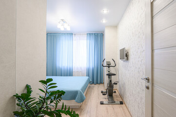 bright, compact room features a bed with blue linens, matching curtains, and an exercise bike. A small TV is mounted on the patterned wall near a white door. A leafy plant adds a touch of nature