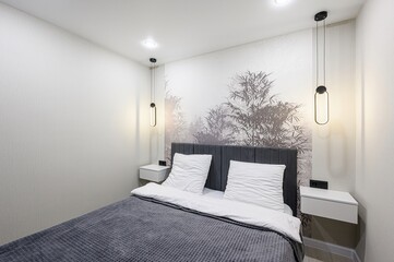 cozy bedroom featuring a bed with grey headboard and blanket, white pillows, bedside tables, and pendant lights. A nature-themed wallpaper adds a serene touch