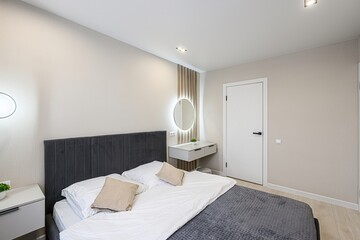 modern bedroom with a gray bed, white linens, beige walls, and a vanity with a round, illuminated mirror. Clean and minimalist aesthetic
