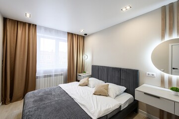 Fototapeta premium modern bedroom featuring a bed with a gray headboard, white linens, brown curtains, and a round, illuminated mirror