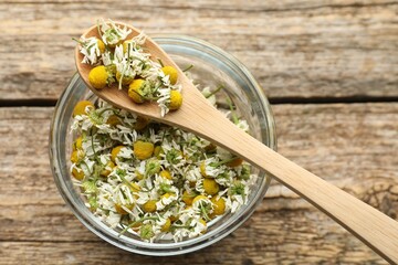 Dry chamomile flowers in bowl and spoon on wooden table, top view