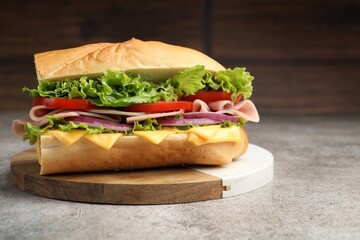 Tasty baguette sandwich with ham, cheese and vegetables on grey textured table against blurred brown background, closeup