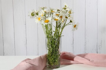 Many beautiful chamomile flowers in vase on white table near wooden wall, closeup