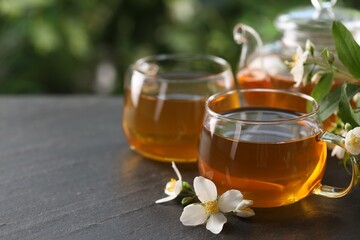 Tasty jasmine tea in cups, teapot and flowers on black table, closeup. Space for text