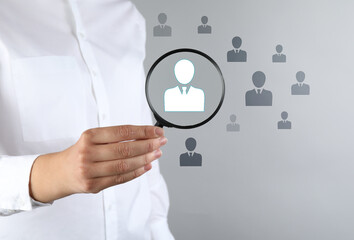 Employee search. HR manager holding magnifying glass over human icons against grey background, closeup