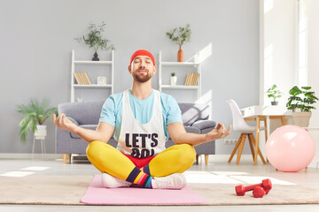 Relaxed peaceful funny young man doing yoga exercise sitting on the floor in lotus pose. Caucasian person with closed eyes meditating on fitness mat in the living room. Healthy lifestyle.
