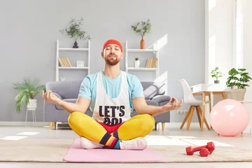 Fotobehang Lotusbloem Relaxed peaceful funny young man doing yoga exercise sitting on the floor in lotus pose. Caucasian person with closed eyes meditating on fitness mat in the living room. Healthy lifestyle.  © Studio Romantic