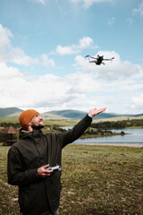 Smiling man reaching for a drone in mid-air above a lake in Zlatibor, Serbia. Beautiful spring mountains and blue sky behind. Travel blogger making video outdoor