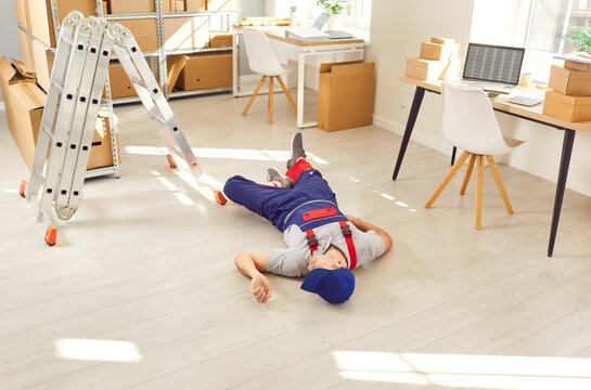 Unconscious male worker storekeeper lying on the floor in overall in warehouse full of cardboard boxes after accident on workplace falling from a ladder. Health insurance and accident at work concept
