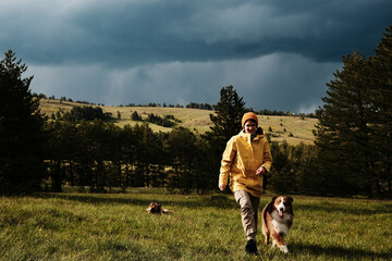Woman in a yellow jacket and her Australian Shepherd walking across a meadow with a forest and hills under a moody sky in Zlatibor, Serbia. Female owner traveling with pets outdoor