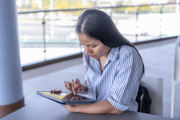 Young Asian woman working on tablet with stylus, focused and productive in a modern indoor/outdoor...