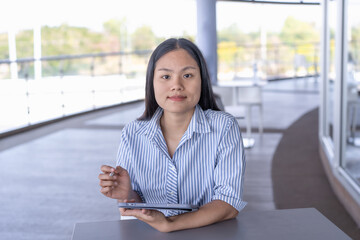 Young Asian woman working on tablet with stylus, focused and productive in a modern indoor/outdoor...