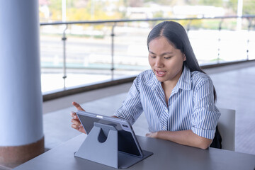 Young Asian woman working on tablet with stylus, focused and productive in a modern indoor/outdoor...