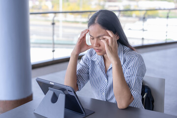 Anxious Asian woman experiencing a headache or stress while working on her tablet, capturing the challenges of remote work and mental strain in a contemporary setting
