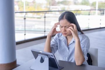 Anxious Asian woman experiencing a headache or stress while working on her tablet, capturing the challenges of remote work and mental strain in a contemporary setting