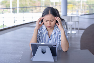Anxious Asian woman experiencing a headache or stress while working on her tablet, capturing the challenges of remote work and mental strain in a contemporary setting