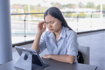 Anxious Asian woman experiencing a headache or stress while working on her tablet, capturing the challenges of remote work and mental strain in a contemporary setting