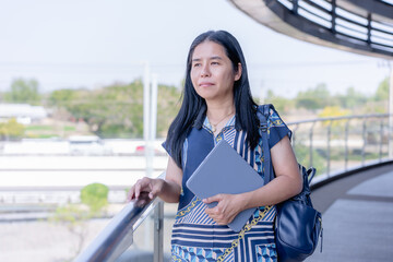 Thoughtful Asian woman standing with a tablet and bag, looking out into a modern, bright open space. Perfect for themes of contemplation, professional lifestyle, and urban environments.