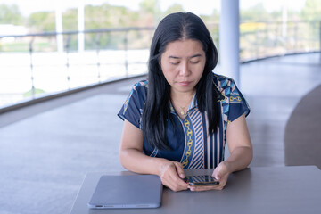 Happy Asian woman engaging with her smartphone at a desk, with a laptop nearby, in a bright, modern indoor-outdoor setting. Ideal for themes of casual tech use, communication, and modern lifestyle