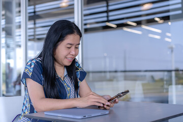 Happy Asian woman engaging with her smartphone at a desk, with a laptop nearby, in a bright, modern indoor-outdoor setting. Ideal for themes of casual tech use, communication, and modern lifestyle