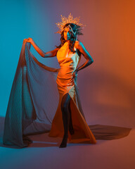 Full length portrait of a female model wearing a  fantasy crown, yellow gown and flowing fabric cape.  Walking pose on studio background with warm golden sunset  lighting