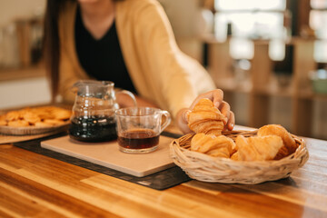 Close-Up of A woman's hand reaches for a golden croissant from a basket, paired with a glass of black coffee on a wooden kitchen counter