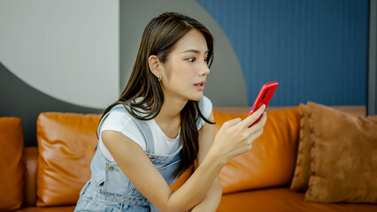 Happy asian young woman sit on sofa at home typing on mobile phone receive great news celebrating...
