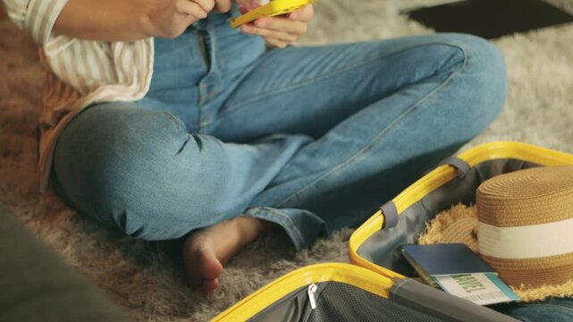 Young woman cross-legged on carpet, packing yellow suitcase with summer travel gear, browsing smartphone, preparing for upcoming adventure with concentrated energy.Traveling and vacation concept - Powered by Adobe