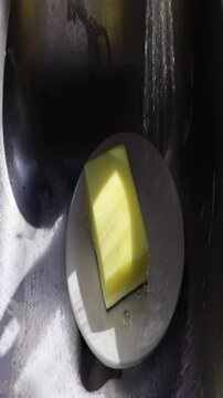 Un primer plano de una esponja amarilla llena de jab&oacute;n liquido que le cae agua en un plato de color blanco dentro de un fregadero. Video vertical