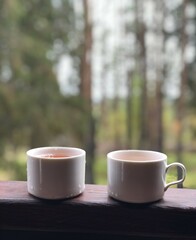 Two white ceramic cups with hot tea overlooking forest view.