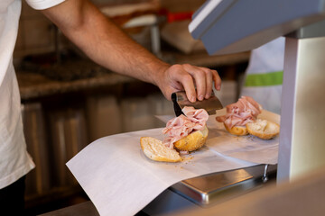 A deli worker slices and arranges cooked ham on fresh rolls, on a countertop in a deli.