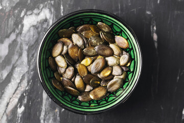 Pumpkin seeds in green bowl. Healthy snack rich in nutrients. Organic roasted pepitas. Top view of crunchy seeds on dark marble background. Natural vegetarian superfood closeup.