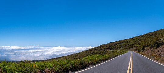 Road winds through lush landscape, with clouds below and blue sky above.