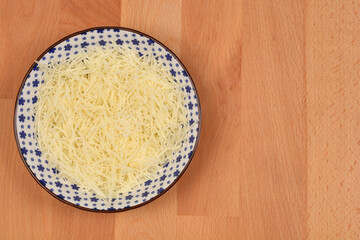 A full bowl of finely grated cheese, presented in a blue and white patterned dish, sits on a warm wooden surface with ample copy space
