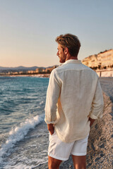 Stylish man in linen shirt standing on beach at sunset, looking at the sea, enjoying a peaceful moment along the scenic coastal shoreline.
