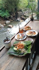 Scenic Outdoor Dining: A Delicious Spread of Indonesian Nasi Uduk and Fried Foods Overlooking a Gentle Stream