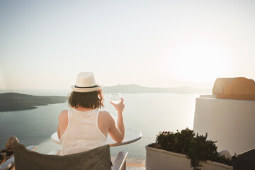 A woman enjoys a scenic sunset view over the Aegean Sea from a Santorini terrace with a glass of wine.