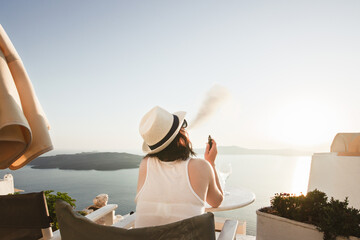 A woman enjoys a scenic sunset view over the Aegean Sea from a Santorini terrace with a glass of wine and a vape.
