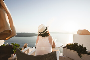 A woman enjoys a scenic sunset view over the Aegean Sea from a Santorini terrace with a glass of wine and a vape.