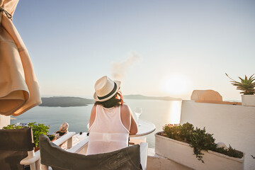 A woman enjoys a scenic sunset view over the Aegean Sea from a Santorini terrace with a glass of wine and a vape.