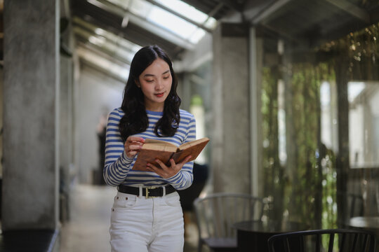 Young woman reading a book while walking in a modern library