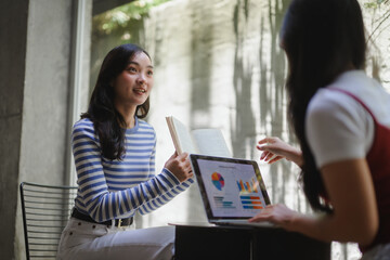 Two young businesswomen discussing marketing strategy using laptop and book