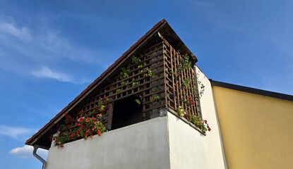 Upper balcony with wooden trellis supports climbing plants and pink and red flowers, set on a white and yellow house with tiled roof under a clear blue sky.