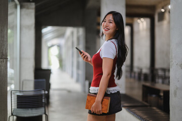 Young student walking in university corridor using smartphone and holding textbook