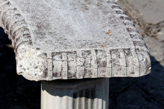 Close-up of an old stone bench with a rough, weathered surface and carved edge pattern, revealing aggregate and signs of age, set outdoors in natural sunlight.